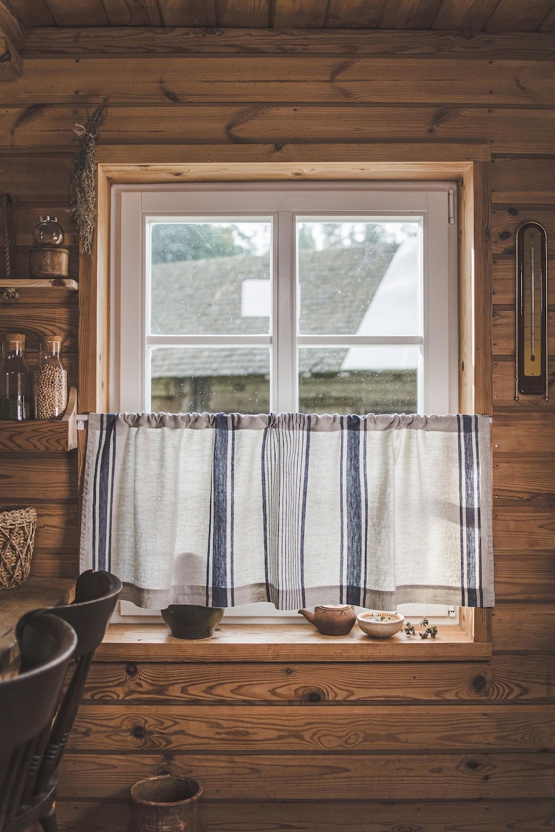 May include: A window with a white frame and a white and blue striped curtain. The window is in a wooden cabin with a wooden wall and a wooden ceiling. There are some jars on a shelf to the left of the window.