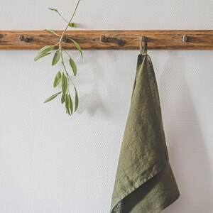 May include: An olive green kitchen towel with a hanging loop suspended from a wooden peg rail. Next to the towel, a sprig of fresh green sage leaves is also hanging from the same wooden peg rail, which features a natural wood grain finish.