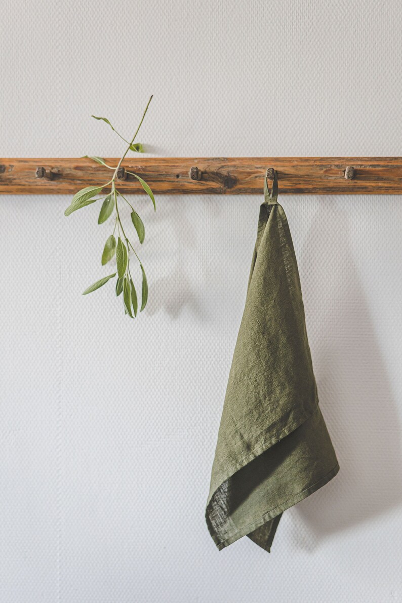 May include: An olive green kitchen towel with a hanging loop suspended from a wooden peg rail. Next to the towel, a sprig of fresh green sage leaves is also hanging from the same wooden peg rail, which features a natural wood grain finish.