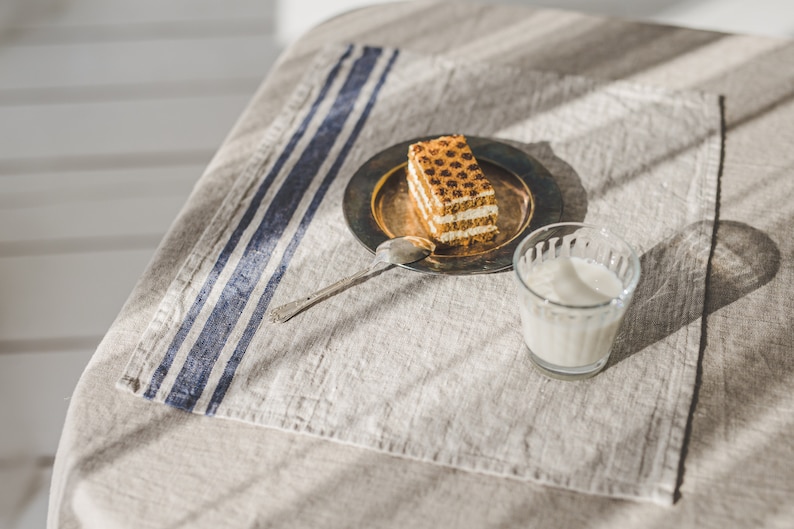 May include: A close-up of a piece of layered cake on a silver plate with a spoon, a glass of milk, and a blue and white striped linen napkin on a beige tablecloth.