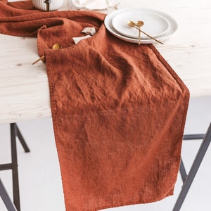 May include: A rust-colored linen table runner with a white ceramic vase and two gold spoons on a white plate.
