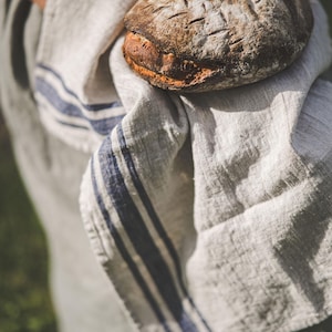 Puede incluir: Una persona sosteniendo un pan reci&eacute;n horneado envuelto en una toalla de cocina de lino a rayas blancas y azules.