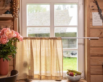 May include: A rustic kitchen scene with a window framed by light wood paneling. A sheer, beige curtain hangs in front of the window. A vase of pink flowers and a bowl of fruit sit on the windowsill. Dried onions and herbs hang nearby.