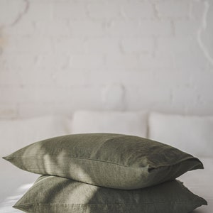 May include: Two olive green linen pillowcases stacked on a white bed. The pillowcases are made of a soft, natural fabric and have a subtle texture.