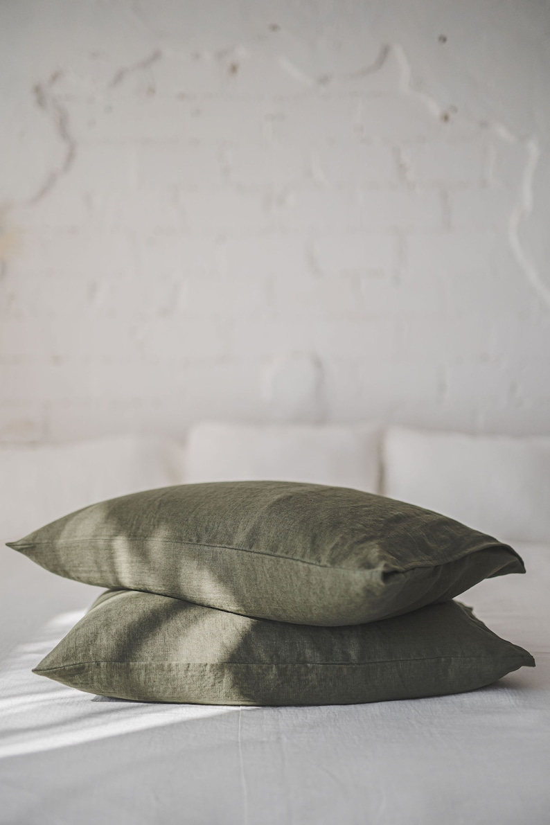 May include: Two olive green linen pillowcases stacked on a white bed. The pillowcases are made of a soft, natural fabric and have a subtle texture.