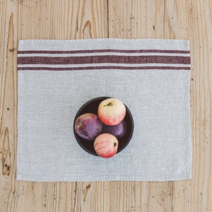 May include: A bowl of red and purple apples on a striped linen placemat. The bowl is black and the apples are arranged in a cluster.