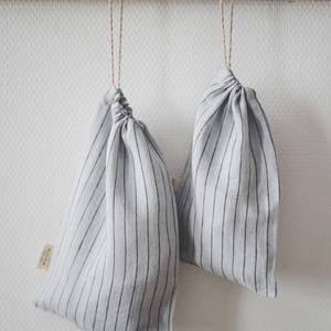 May include: Two gray and white striped fabric bags hanging from a wooden rack. The bags are tied at the top with twine and have a small tag attached. The bags are hanging against a white wall.