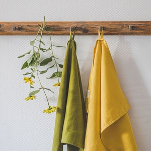 May include: Two rectangular kitchen towels, one in olive green and one in mustard yellow, each featuring a small fabric hanging loop at the top. The towels are suspended from a wooden peg rail alongside a stem of yellow flowers with green leaves.