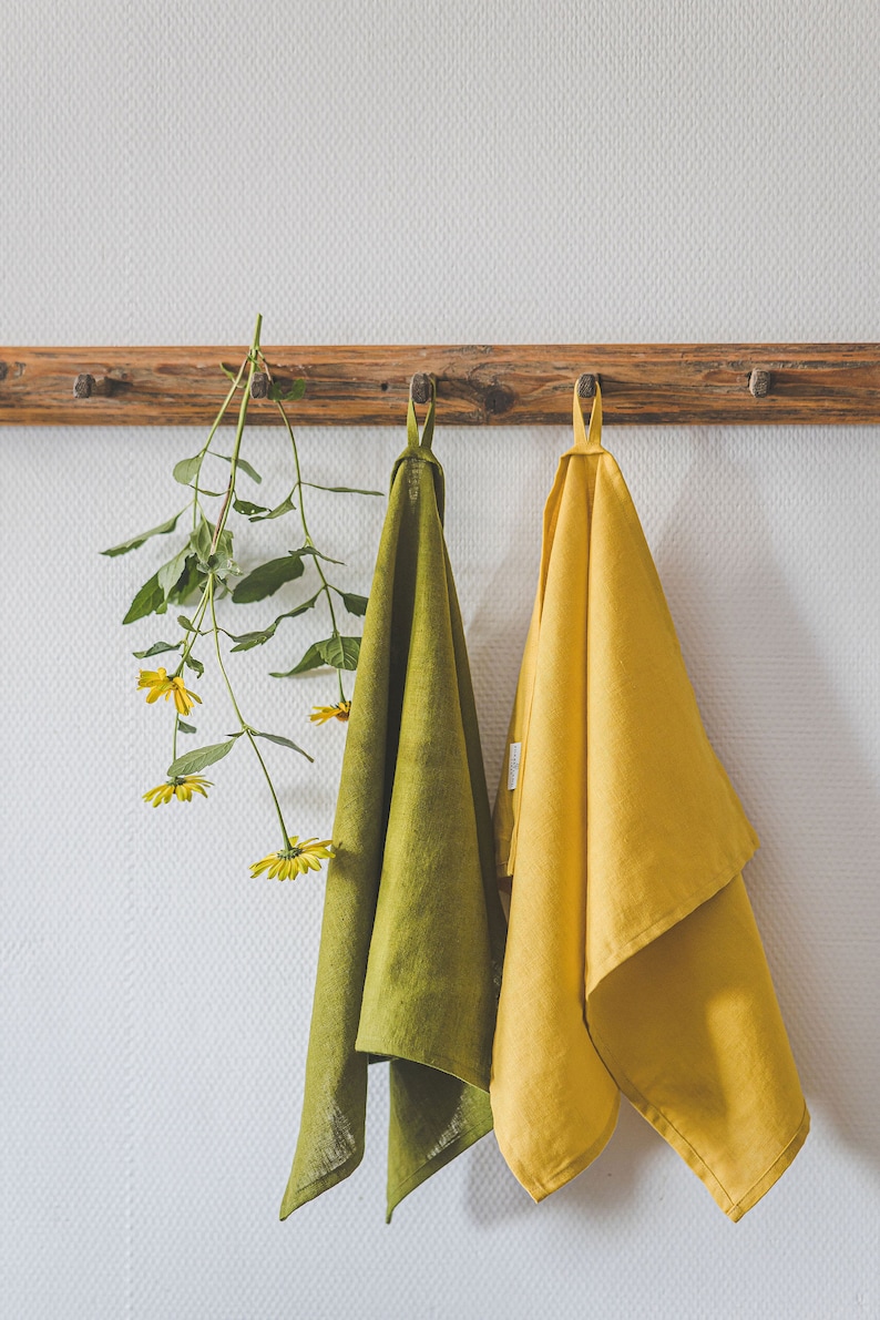May include: Two rectangular kitchen towels, one in olive green and one in mustard yellow, each featuring a small fabric hanging loop at the top. The towels are suspended from a wooden peg rail alongside a stem of yellow flowers with green leaves.