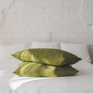 May include: Two green linen pillowcases stacked on a white bed. The pillowcases are made of a soft, natural fabric and have a subtle sheen.