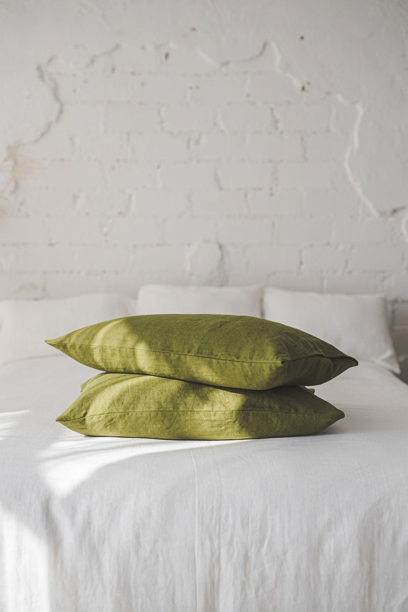 May include: Two green linen pillowcases stacked on a white bed. The pillowcases are made of a soft, natural fabric and have a subtle sheen.