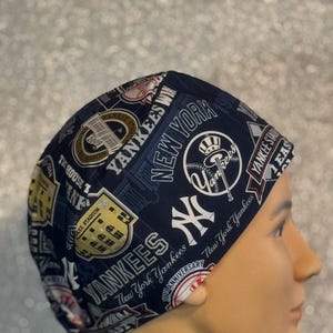 May include: A navy blue Yankees cap featuring various logos, text, and the words "New York Yankees." The cap has a repeating pattern of team-related imagery, including the team's logo and stadium details.