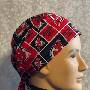 May include: A red, black, and white New Jersey Devils patterned head wrap. The head wrap features the team logo and the words "New Jersey Devils" in a repeating pattern. The head wrap is tied at the back.