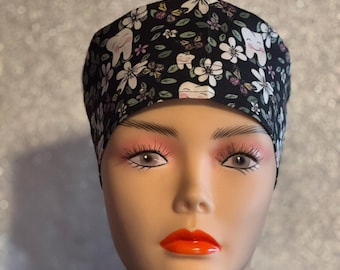 May include: Black scrub hat with a white floral and tooth pattern. The hat has a tie on the back.
