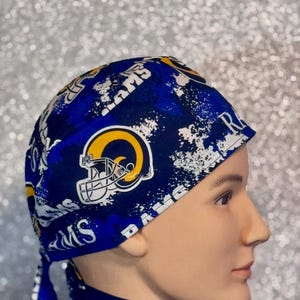 May include: Blue and white patterned head covering featuring the Los Angeles Rams logo. The head covering has a tie in the back and is made of a soft, flexible material. The Rams logo is a yellow ram's head inside a blue circle.