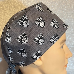 May include: A gray and black patterned surgical scrub hat with a tie. The hat features a repeating pattern of the Brooklyn Nets logo and the words "Nets Brooklyn".