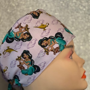 May include: A light purple scrub cap featuring a repeating pattern of a cartoon princess, genie lamps, and clouds. The princess is wearing a teal outfit and has black hair. The cap is tied at the back.