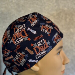 May include: A blue and orange patterned surgical scrub hat with the text "Best Dad Ever!" repeated throughout the design. The hat has ties to secure it in place.