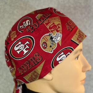 May include: Red and gold patterned surgical cap with the San Francisco 49ers logo. The cap has a tie at the back.