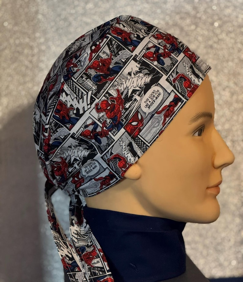 May include: A white and red surgical scrub hat with a comic book pattern featuring Spiderman. The hat has a tie that is tied around the back of the head.