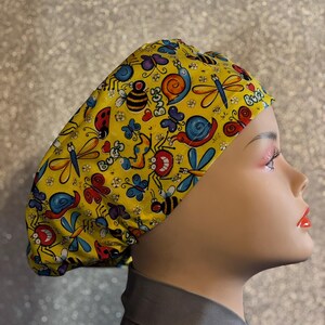 May include: Yellow scrub hat with a colorful bug print. The print includes ladybugs, butterflies, bees, snails, and dragonflies. The hat has a tie at the back.