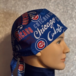 May include: A blue Chicago Cubs bandana-style head covering. The fabric is patterned with the team's name and logo in white and red. The head covering is tied at the back, and is displayed on a mannequin head.