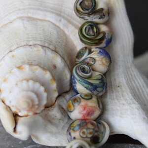 May include: A strand of colourful glass beads with a swirling pattern, strung on a black cord. The beads are arranged in a row and are resting on a white seashell.