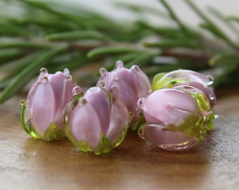 May include: Five pink glass beads shaped like tulips with green accents. The beads are arranged on a wooden surface.