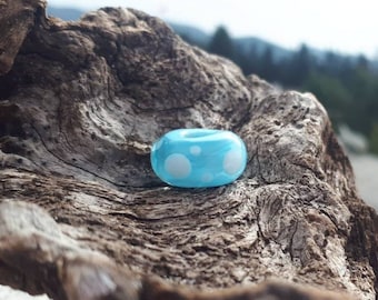 May include: A light blue glass bead with white polka dots. The bead is sitting on a piece of driftwood.