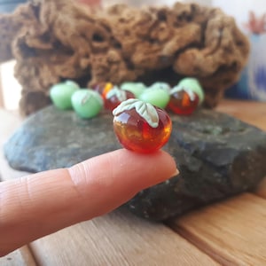 Pode incluir: Uma pequena maçã de vidro translúcido vermelho e laranja com uma folha verde está em cima de um dedo. Várias maçãs de vidro verdes e vermelhas estão no fundo, sugerindo um item decorativo ou artesanal com tema de frutas.