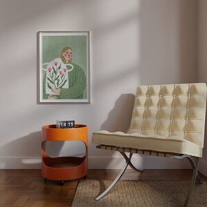 May include: A framed illustration of a person holding a bouquet of pink flowers. The illustration is set against a green background. A clock displaying 14:15 is on a small orange side table. A white chair with a tufted back is in the foreground.