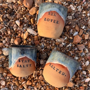 May include: Three handcrafted ceramic mugs with a seaside theme. Each mug features a tan base, a blue and white glazed top, and a unique phrase: "SEA LOVER," "STAY SALTY," and "SALTED." The mugs are presented on a bed of small, colourful pebbles.