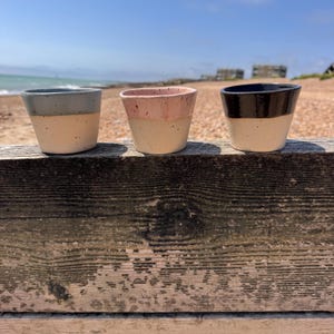 May include: Three small ceramic cups with a speckled beige base and coloured rims in grey, pink, and black. The cups are displayed on a weathered wooden surface with a beach and sea in the background.