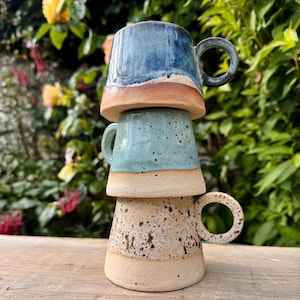 May include: Three handmade ceramic mugs stacked. The top mug is blue, the middle is turquoise, and the bottom is speckled beige. Each mug has a rounded handle and a light-coloured base. The mugs are on a wooden surface.