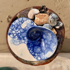May include: A white ceramic dish with a blue bubble design. The dish is shaped like a shell and has a small dip in the centre. The dish is sitting on a brown surface with some stones.