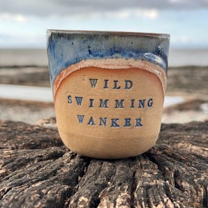 May include: A ceramic mug with a tan base and a blue glazed rim. The text WILD SWIMMING WANKER is stamped in dark blue letters on the front. The mug has a smooth, rounded shape and a textured, earthy appearance, contrasting the blue glaze.