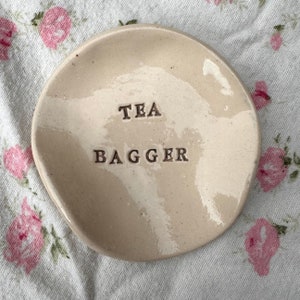 Op de afbeelding: Een witte keramische theezakjeshouder met de tekst "TEA BAGGER" erop.
