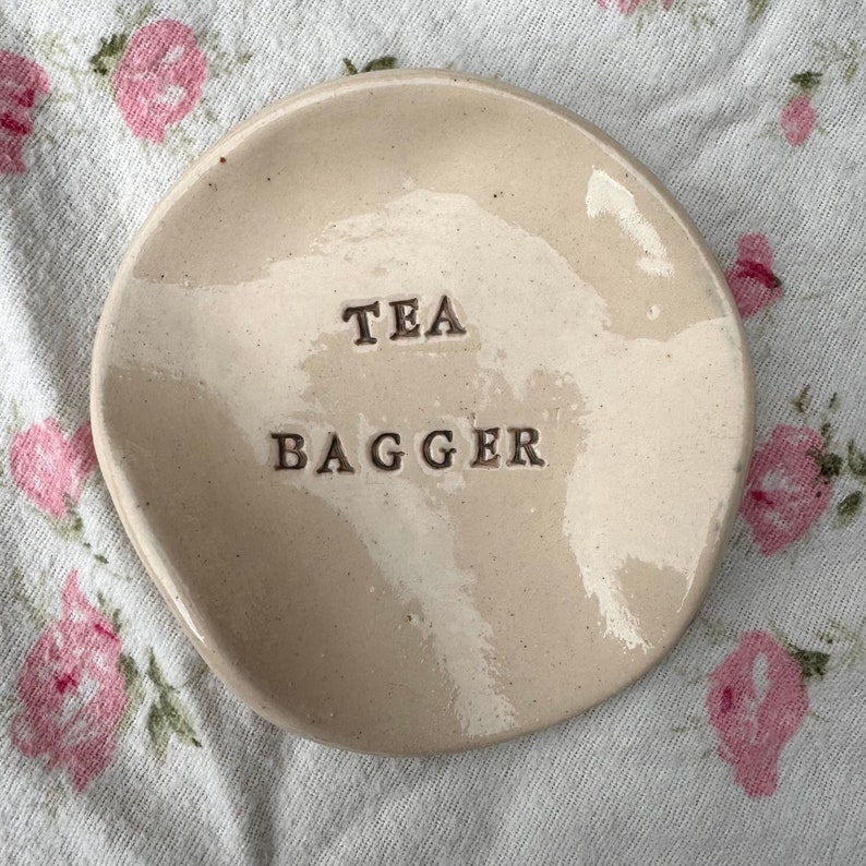 Op de afbeelding: Een witte keramische theezakjeshouder met de tekst "TEA BAGGER" erop.
