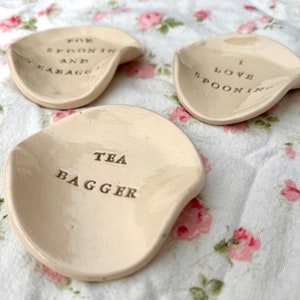 Op de afbeelding: Drie keramische lepelsteunen met verschillende spreuken. De eerste zegt "Voor lepel en theezakje". De tweede zegt "Ik hou van knuffelen". De derde zegt "Tea bagger".