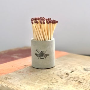 May include: A grey concrete matchstick holder with a bee design. The holder is filled with wooden matches with brown tips.