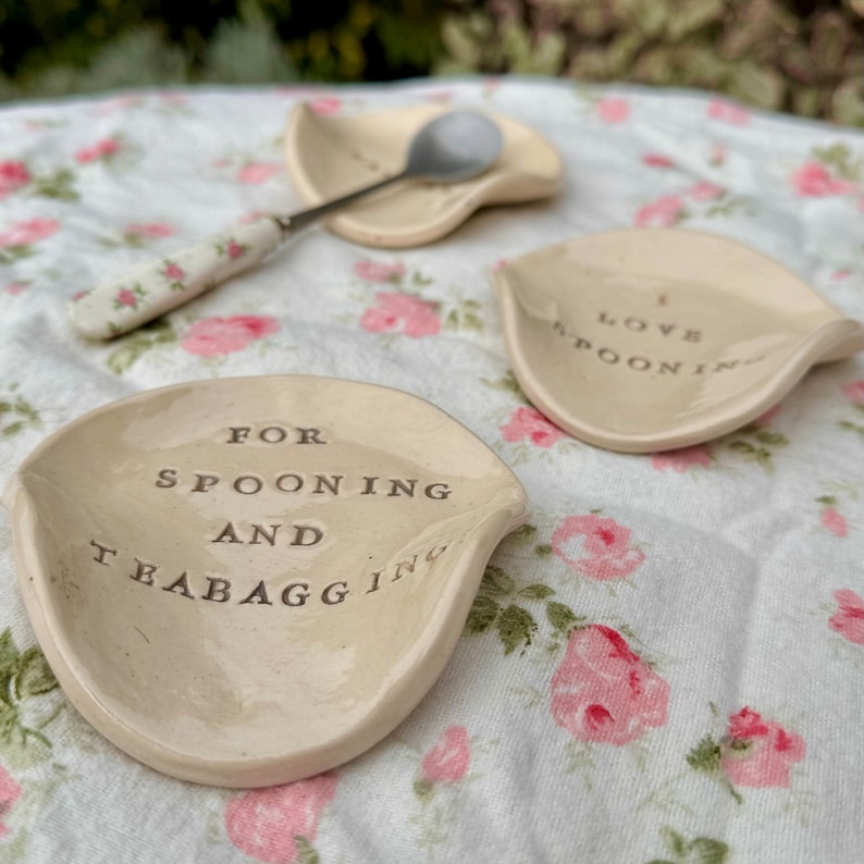 Op de afbeelding: Drie keramische lepelsteunen met de tekst "For Spooning and Teabagging", "I Love Spooning" en "I Love Spooning" op een bloemenkleed.
