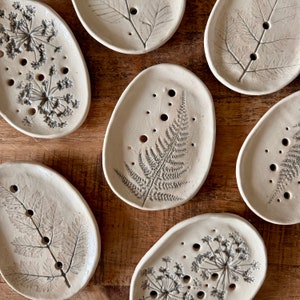 May include: Set of six ceramic soap dishes featuring various botanical designs. Each dish showcases a different plant motif, including ferns, leaves, and flowers. The dishes are white with grey accents and have a rustic, handmade appearance.