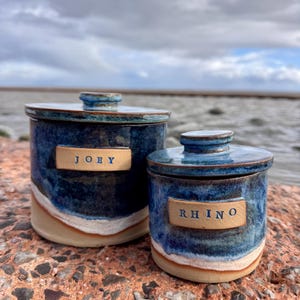 May include: Two cylindrical ceramic canisters with lids, featuring a deep blue glaze that transitions into a white band and a natural tan base. The larger canister has a rectangular label reading JOEY, and the smaller one reads RHINO.