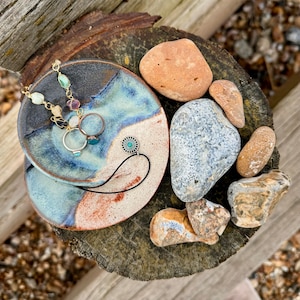 May include: A ceramic dish with a blue and white glaze, featuring a gold necklace with colourful beads, two rings, and a pendant with a turquoise stone. The dish is surrounded by various smooth, grey and brown stones.