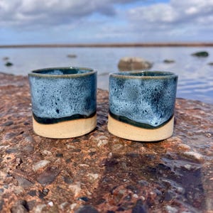 May include: Two cylindrical ceramic cups featuring a speckled blue and dark teal glaze on the upper portion and a smooth, unglazed beige base. The cups have straight sides and a simple, handleless design with a distinct colour transition.