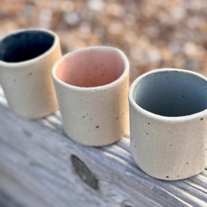 Puede incluir: Tres tazas de cerámica hechas a mano con exteriores moteados de color blanquecino. Los interiores están coloreados en tonos azul oscuro, rosa y azul claro. Las tazas son cilíndricas y se asientan sobre una superficie de madera, mostrando una estética rústica.