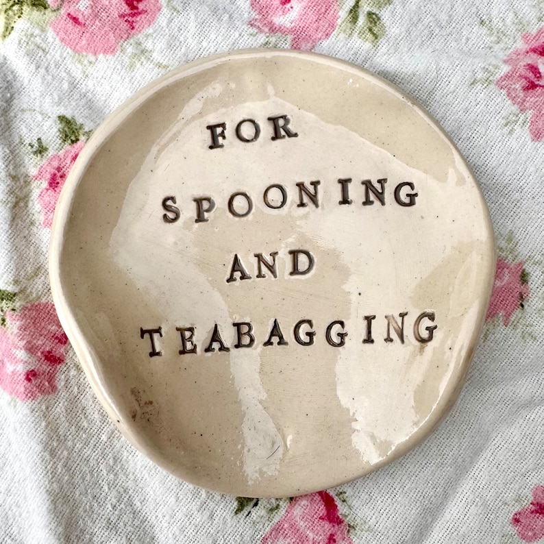 Op de afbeelding: Een wit keramisch schaaltje met een licht onregelmatige vorm. Het schaaltje heeft de tekst "FOR SPOONING AND TEABAGGING" in het zwart erop.