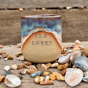 May include: A ceramic tumbler with a coastal design. The top rim has a blue and brown glaze, leading to a tan midsection with the words "SEA LOVER" etched. The tumbler is surrounded by pebbles and shells, evoking a seaside feel.