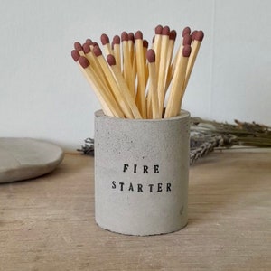 May include: A grey concrete match holder with the words "FIRE STARTER" printed on the front. The holder is filled with wooden matches with red tips.