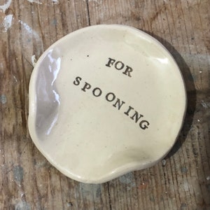 Op de afbeelding: Een wit keramisch schaaltje met een licht onregelmatige vorm. Het schaaltje heeft de woorden "FOR SPOONING" erop geschreven in gouden letters.